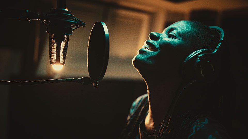 Female vocalist wearing headphones and singing into microphone in recording studio.