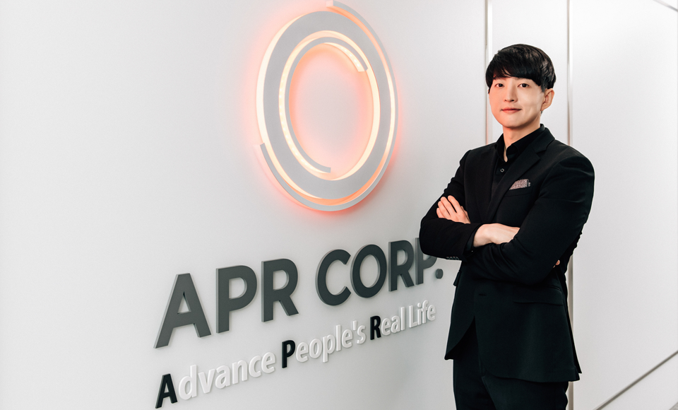 صورة للرئيس التنفيذي لشركة APR، بيونغ هون كيم، يرتدي بدلة سوداء ويقف وذراعاه متقاطعتان بجانب صورة شركة APR وشعارها «تحسين حياة الناس الحقيقية» المعلق على جدار المكتب.