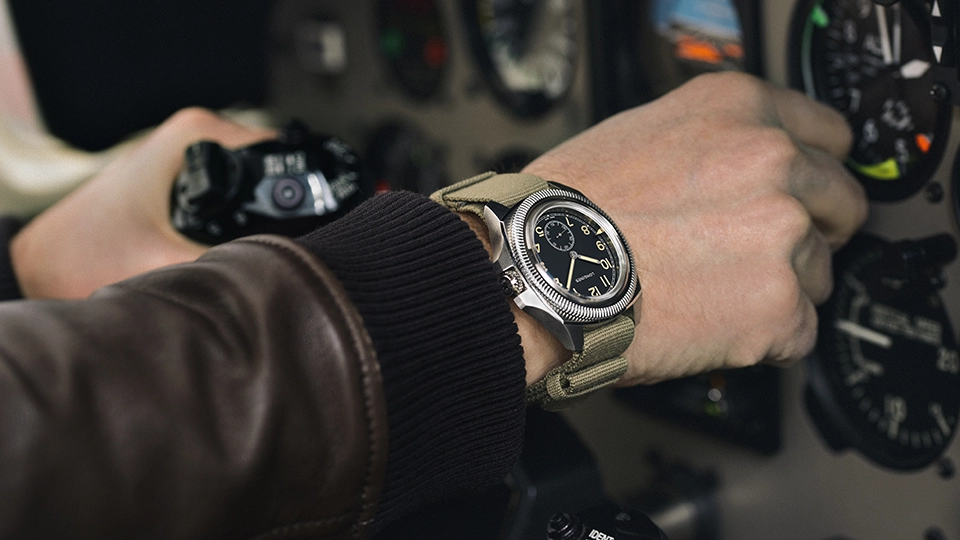 A pilot's wrist wearing an aviator chronograph watch with black dial and beige NATO strap, shown while gripping aircraft controls in a cockpit. 
