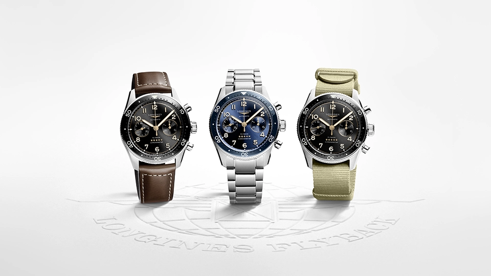Three Longines aviator chronograph watches displayed side by side on a white surface with an embossed circular logo beneath them. From left to right: a watch with black dial and brown leather strap with contrast stitching; a watch with blue dial and stainless steel bracelet; and a watch with black dial and beige NATO-style fabric strap. All three feature bidirectional rotating bezels, Arabic numeral hour markers, chronograph subdials, and polished steel cases in a classic aviation-inspired design