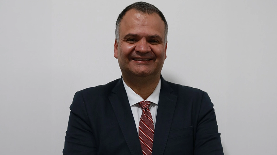 A man in a dark suit with a white shirt and red striped tie smiles at the camera, standing against a plain white background.